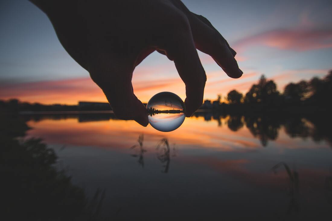 Tussen duim en wijsvinger hangt een glazen knikker die een waterkant tegen avondlucht uitvergroot. Het suggereert een surrealistisch verhaal.